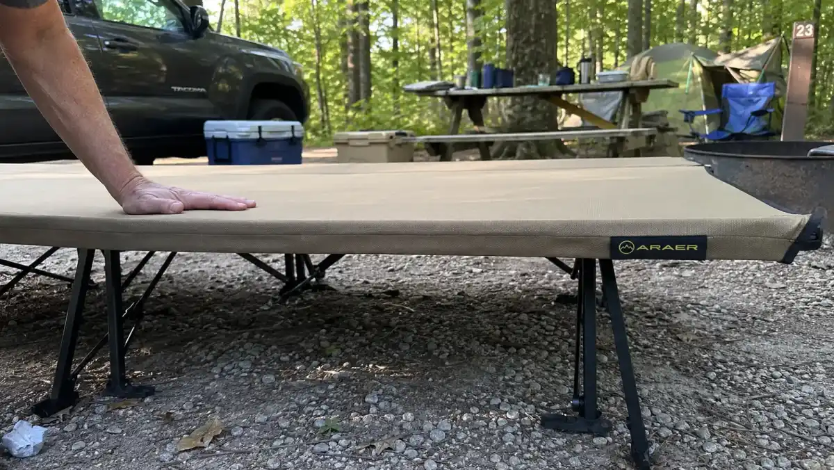 Close-up of a hand pressing on the fabric deck of the ARAER camping cot showing the gap underneath between the fabric and the ground